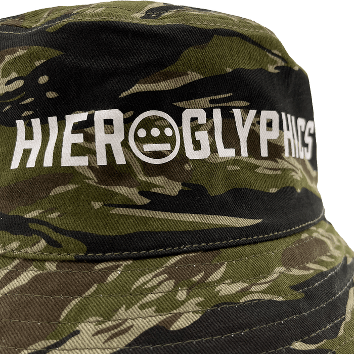 A close-up view of the Hiero Reversible Camo Bucket hat by Hieroglyphics, showcasing the word "Hieroglyphics" in white, uppercase letters. The text features a unique circle design with three horizontal lines and two dots forming a face-like symbol in place of the letter "O.