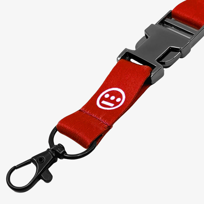 A close-up image of the Hiero VIP Lanyard 2.0 by Hieroglyphics, showcasing a red lanyard with a white smiley face featuring a neutral expression. The lanyard includes a black plastic buckle and a metal swivel hook at the end. The partially unclasped buckle highlights the texture and design of both the fabric and hardware.