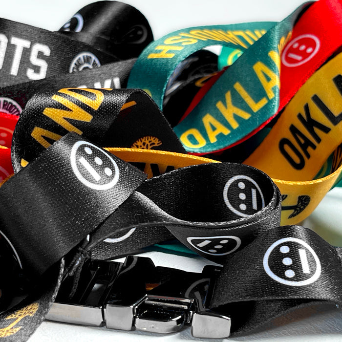 A vibrant display of Hiero VIP Lanyard 2.0 from Hieroglyphics features various hip-hop designs, including text like "OAKLAND" and distinctive logos. The lanyards come in black, yellow, red, and green. Intertwined together, a metal clasp is prominently visible at the forefront, showcasing their texture and vivid colors in a close-up shot.