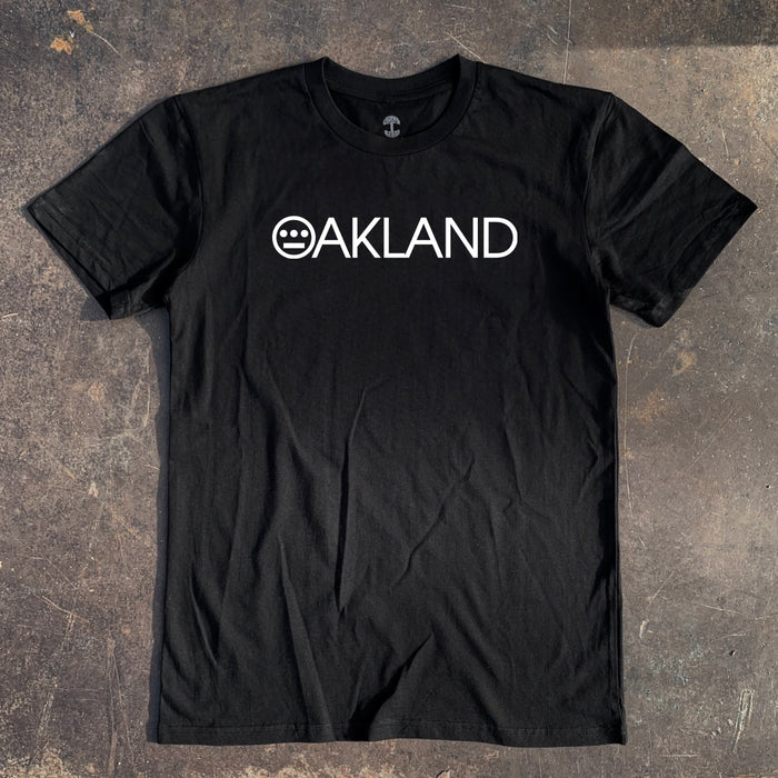 The Hieroakland Tee by Hieroglyphics, a black, 100% cotton T-shirt with a classic fit, lies on a rustic, worn surface. The shirt features "OAKLAND" in white capital letters across the chest with the "O" stylized as a smiley-face graphic, creating a unique design element perfect for Town business.