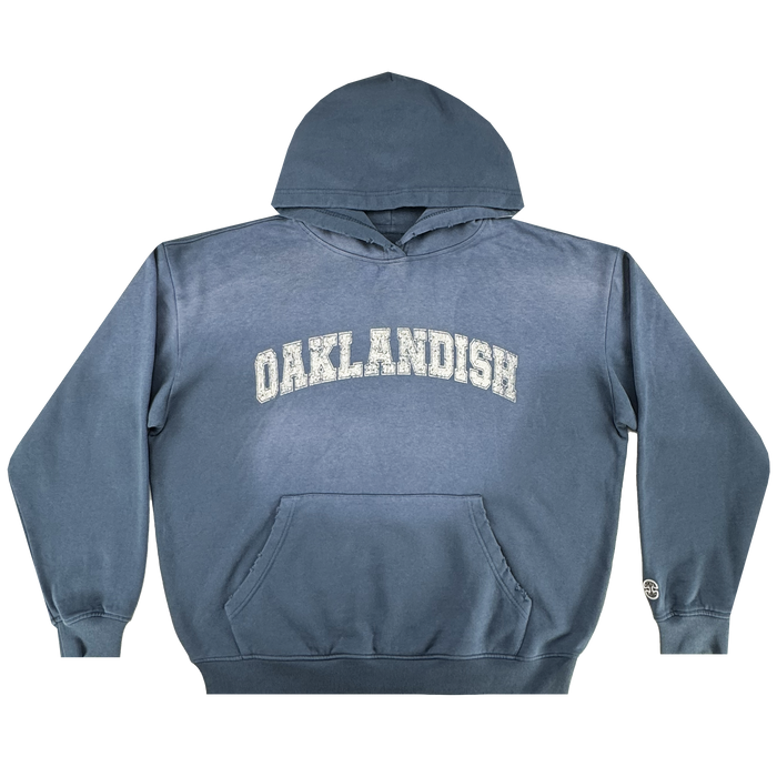 The Homage Hoodie by Oaklandish is a blue 100% cotton pullover with a front pocket, "OAKLANDISH" in large white distressed letters on the chest, and a small round logo on the left cuff—perfect men's outerwear for everyday style.