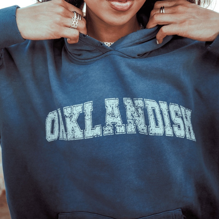 A person tugs at the collar of an oversized blue Oaklandish Homage Hoodie, showing off bold, distressed "OAKLANDISH" lettering across the chest. Only their hands with rings and the lower half of their face are visible.