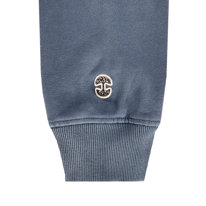 A close-up of the Oaklandish Homage Hoodie’s blue ribbed cuff, made from 100% cotton, shows a small embroidered black and white circular patch with a stylized tree logo near the hem.