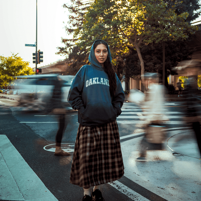 A woman stands confidently on a city street corner in an Oaklandish Homage Hoodie, blue and oversized with “OAKLAND,” paired with a plaid skirt. Hands in pockets, she faces the camera as blurred pedestrians pass and sunlight streams through trees behind her.