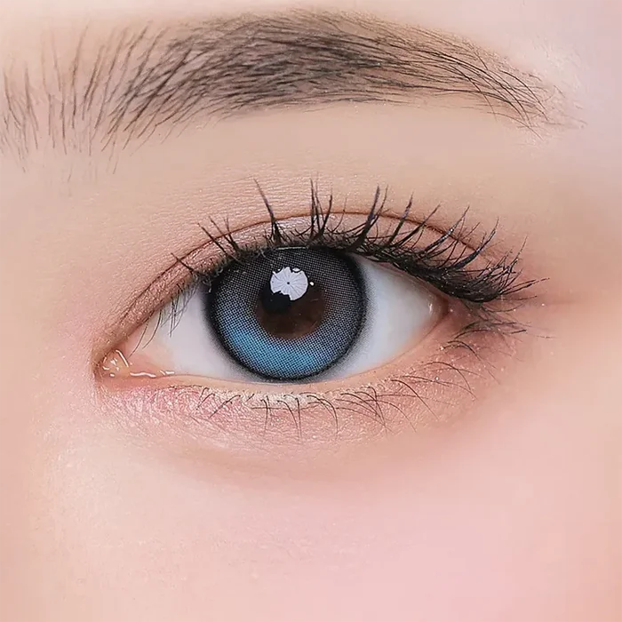 A close-up of a human eye with long lashes wearing i-Girl One Pick Balance Forest Tea Blue contact lenses by i-Girl, framed by natural skin and a neat eyebrow, showcasing the fresh Douyin girl style.