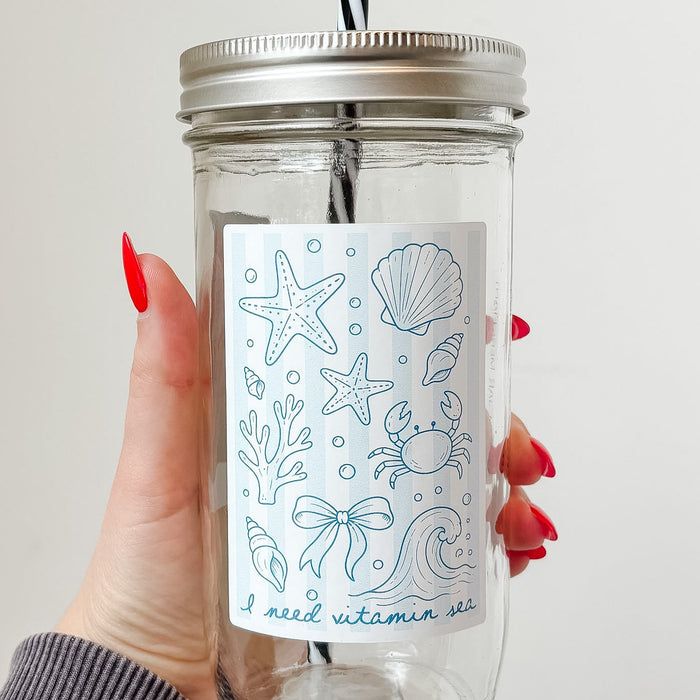 Hand holding mason jar tumbler up close that reads i need vitamin sea with starfish, shell, crab, coral and other ocean designs on pale blue and white striped background