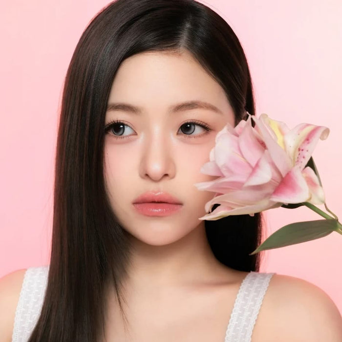 A young woman with long straight dark hair wears i-Sha One Pick Only One 1-Day Velvet Smoke Grey (10pk) contacts, holding a large pink lily near her face. She’s in a white top against a soft pink gradient, looking calm and serene.