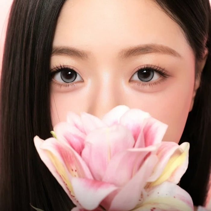 A woman with straight dark hair holds a large pink flower close to her face, partially covering her mouth and nose. Her eyes, enhanced by i-Sha One Pick Only One Velvet Smoke Grey colored contacts from i-Sha, are wide open, and the background is softly blurred in pink tones.