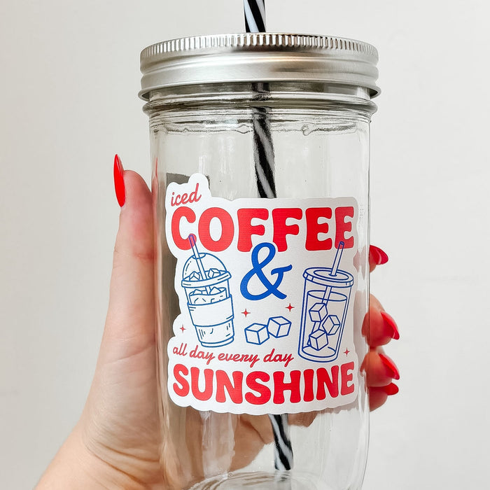 Hand holding mason jar tumbler up close that reads iced coffee & sunshine all day every day in red ink with blue iced drink designs on white background