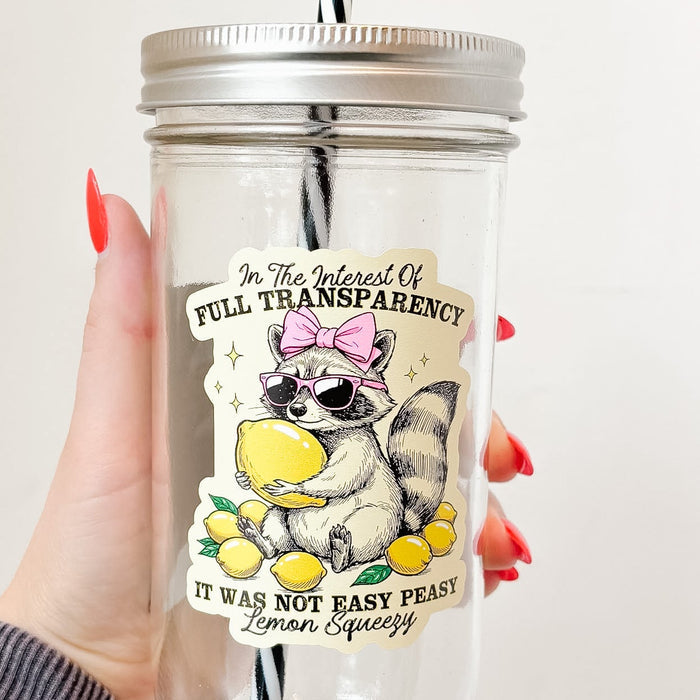 Hand holding mason jar tumbler up close that reads in the interest of full transparency it was not easy peasy lemon squeezy with a raccoon wearing pink sunglasses and bow holding a lemon and surrounded by lemons design on pale yellow background