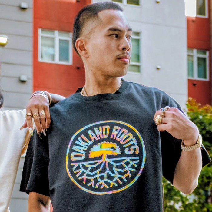 A person stands outdoors by a modern building, looking to the side. They're wearing the Oakland Roots SC Iridescent Tee and pointing to its colorful tree logo. Multiple rings decorate their fingers; another arm is draped over their shoulder.