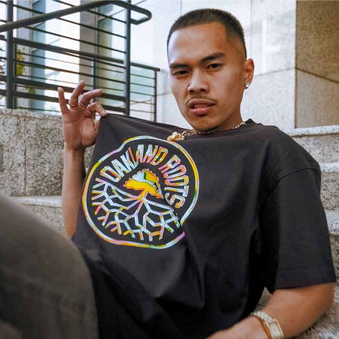 A person with short hair sits on outdoor stone steps, confidently holding up the Iridescent Tee by Oakland Roots SC, featuring a colorful logo. Behind them are metal railings and a modern stone building, reflecting their Oakland Roots pride.