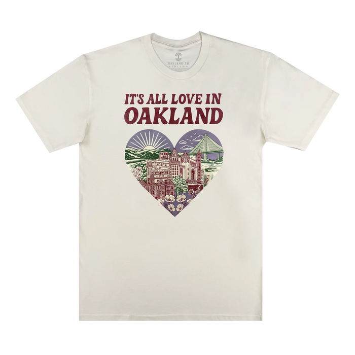 The Oaklandish It's All Love in Oakland Tee is a cream, 100% cotton shirt with bold "IT'S ALL LOVE IN OAKLAND" text above a heart graphic featuring Oakland landmarks, flowers, the sun, and cityscape in green, purple, and red.