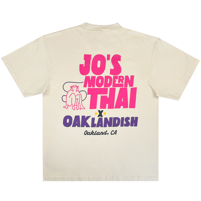 Jo’s Modern Thai X Oaklandish Tee by Other: An off-white heavyweight t-shirt featuring bold pink and purple text—“JO’S MODERN THAI,” “OAKLANDISH,” and “Oakland, CA.” The collaborative design includes a pink line drawing of a dog above “OAKLANDISH” and a small yellow star between the words.