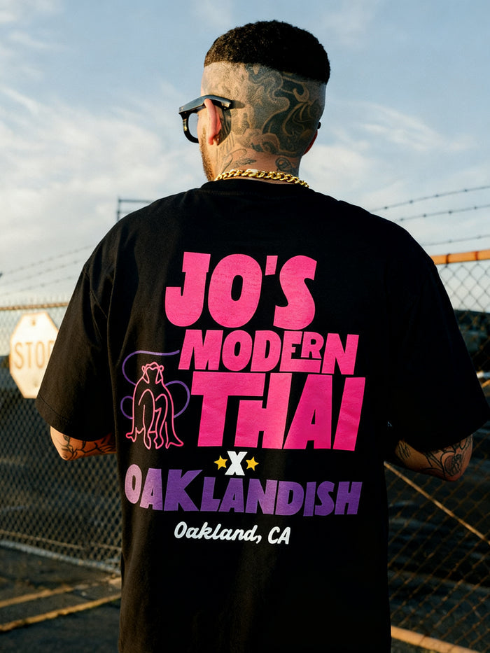 Wearing the Jo’s Modern Thai X Oaklandish Tee by Other, a person with tattoos and sunglasses faces away from the camera. The heavy-weight t-shirt features “JO’S MODERN THAI x OAKLANDISH Oakland, CA” in pink, purple, and yellow lettering, alongside a line drawing of two figures. Outdoors at sunset near a chain-link fence, this collaborative tee makes a bold statement.