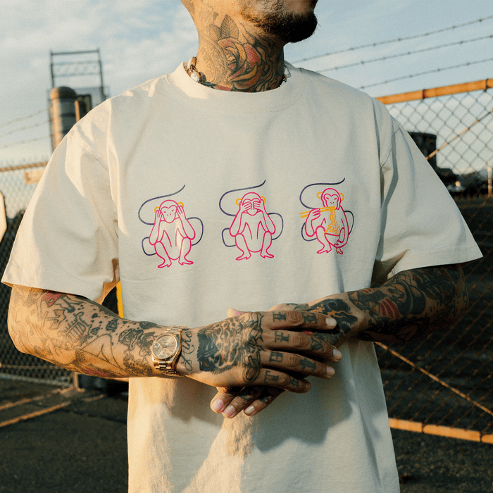 Wearing the Jo’s Modern Thai X Oaklandish Tee by Other, a tattooed individual stands outdoors against an industrial backdrop and a fence. The cream heavyweight t-shirt features three colorful line-drawn monkeys in “see no evil, hear no evil, speak no evil” poses—a striking design from this unique collaboration. A watch adorns their wrist, adding to the look.