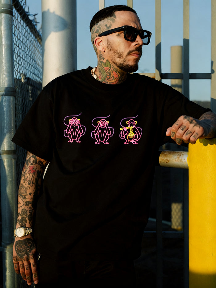 A tattooed man in sunglasses and a heavy-weight Jo’s Modern Thai X Oaklandish Tee by Other—featuring pink and yellow cartoon monkeys—leans on a yellow pole by a metal fence, looking to the side. The collaborative t-shirt displays “see no evil, hear no evil, speak no evil” monkey graphics.