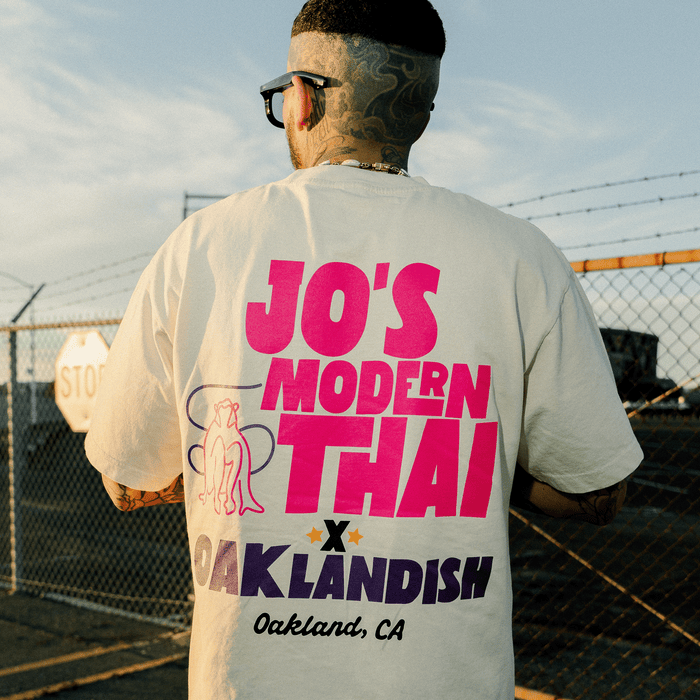 A person with short hair and tattoos, wearing sunglasses and facing away, showcases the Jo’s Modern Thai X Oaklandish Tee by Other. The cream heavyweight t-shirt features bold text— “JO’S MODERN THAI X OAKLANDISH Oakland, CA”— and a small illustration of two figures on the back. A fence and stop sign in the background highlight the standout design of this collaborative piece.