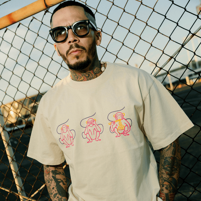 A tattooed man wearing sunglasses and a beaded necklace stands by a chain-link fence, dressed in the Jo’s Modern Thai X Oaklandish Tee from Other—a heavyweight beige shirt adorned with three cartoon monkeys in playful pink and yellow line art. Sunlight casts long shadows in this outdoor scene.