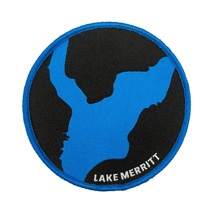 The Oaklandish Lake Merritt Patch is a circular embroidered iron-on patch that features a stylized map outline of Lake Merritt in blue against a black background, with a blue border. The label "LAKE MERRITT" is embroidered in white at the bottom right edge, making it the perfect customized patch for your accessories.