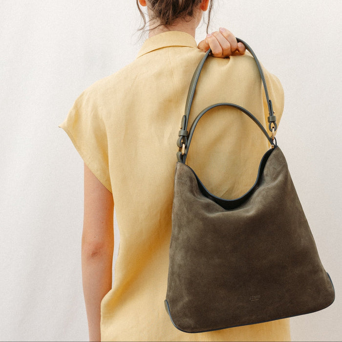 Model holding STOW London's Curve in Dark Green suede featuring the detachable shoulder strap and top handle with gold hardware