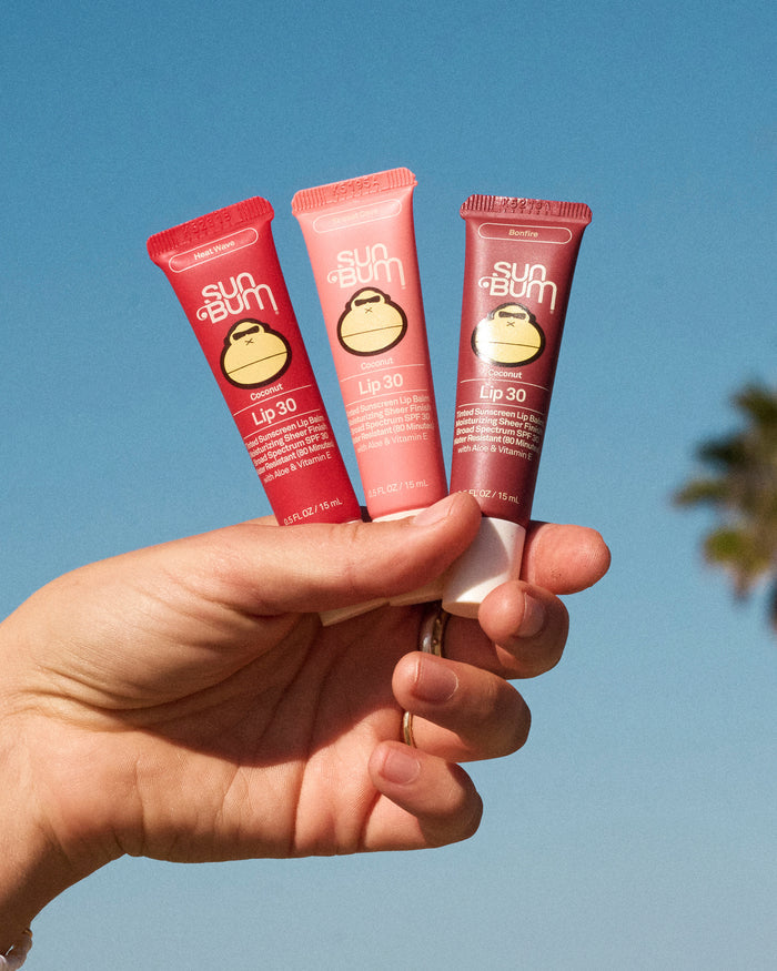 Hand holding three Sun Bum Lip 30 Trio lip balms in red, pink, and brown tubes against a blue sky background