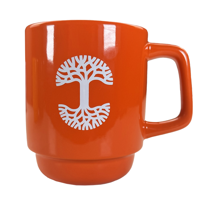 The Oaklandish Logo Stack Mug is an orange ceramic coffee cup with a large handle, featuring a white stylized tree with spreading roots and branches centered on the front. The background is plain white.