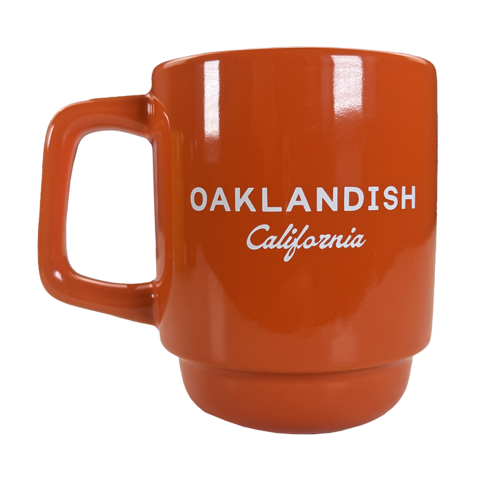 The Oaklandish Logo Stack Mug is an orange ceramic drinkware cup with a squared handle and "OAKLANDISH California" printed in white, shown on a white background. The design is minimal, with no extra decorations or graphics.