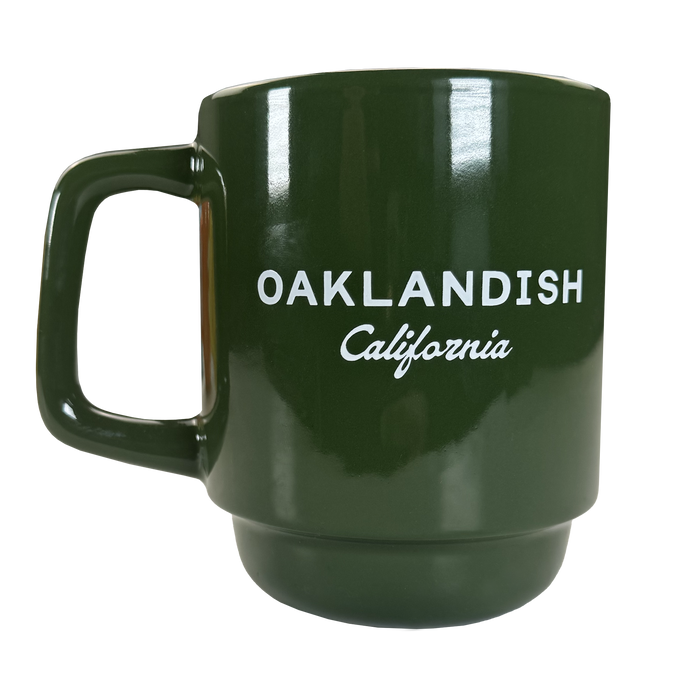 The Oaklandish Logo Stack Mug is a dark green ceramic mug with a large handle, featuring bold white "OAKLANDISH California" text on the front. Its modern design stands out against the plain background.