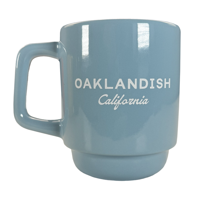The Oaklandish Logo Stack Mug is a light blue ceramic mug with a squared handle, glossy finish, and white "OAKLANDISH California" text. This stackable drinkware piece is shown on a plain white background.
