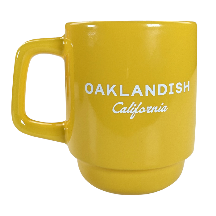 The Logo Stack Mug by Oaklandish is a bright yellow ceramic mug with a large handle. "OAKLANDISH" in bold white uppercase letters and "California" in small cursive are printed on the front. Background is plain white.