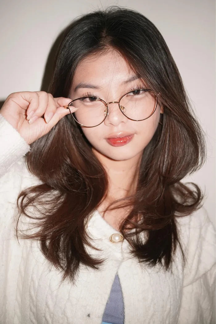 A woman with long dark hair wears EyeCandys Madame Cat Eye lightweight metal frames and a white textured top, looking at the camera while holding her glasses with one hand and slightly smiling.