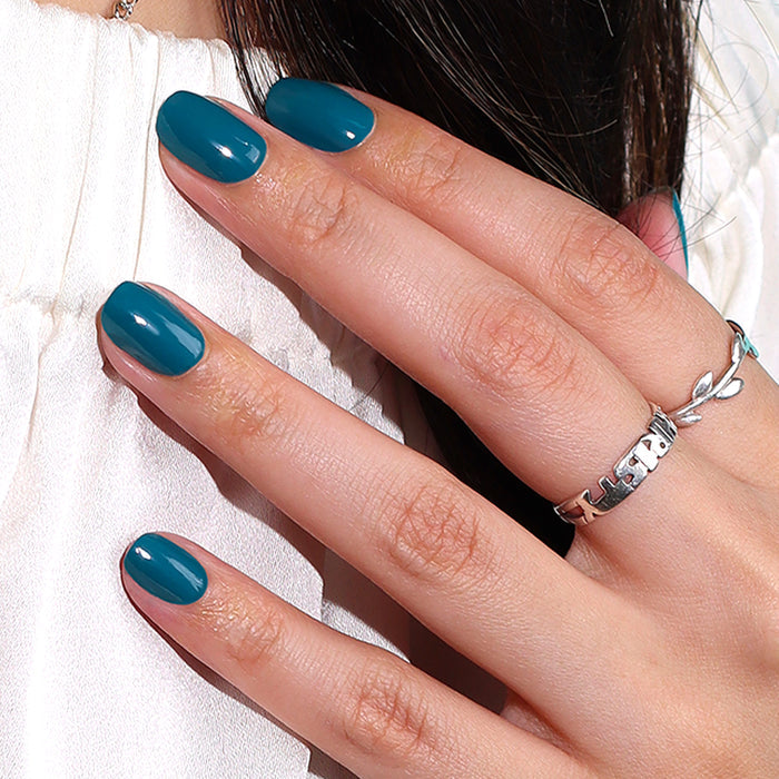 A hand with LONDONTOWN's "Magpie" peacock teal manicured nails and a silver ring rests against a white surface, making a sophisticated statement.