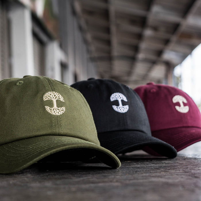 Three Micro Logo Dad Hats from Oaklandish are lined up on a surface. These caps come in olive green, black, and maroon, each featuring adjustable strap backs. The setting appears to be an urban or industrial environment with metal beams and windows in the background, slightly out of focus.