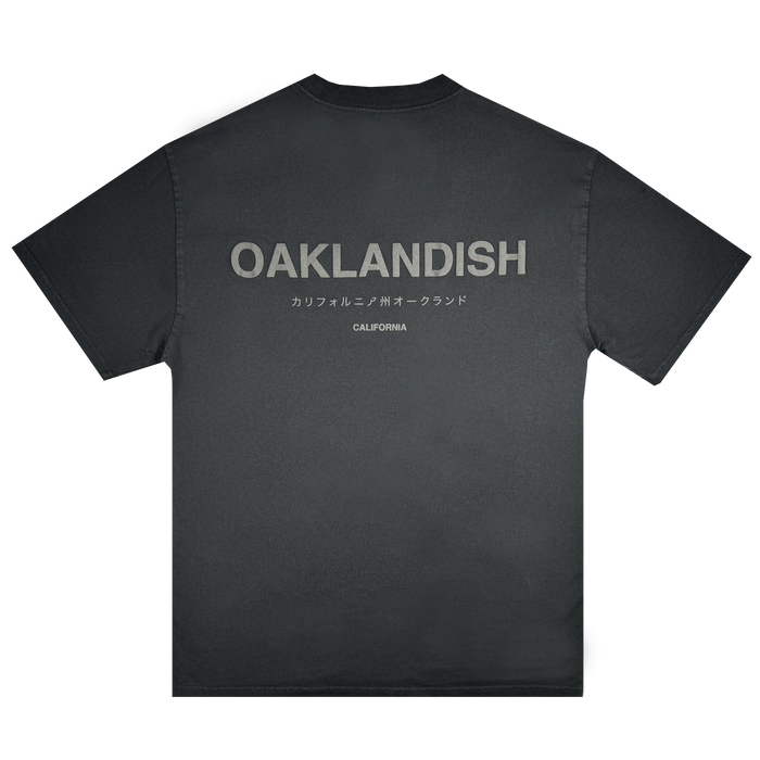 The Modern Type Heavy Tee by Oaklandish, made from heavyweight cotton in a faded black shade, showcases bold gray "OAKLANDISH" lettering across the back. Below it, the word "CALIFORNIA" appears in smaller gray font alongside Japanese text. The shirt is displayed against a plain white background.