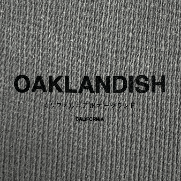 The Modern Type Heavy Tee by Oaklandish showcases a gray fabric with "OAKLANDISH" prominently displayed in bold black letters. Below it, Japanese characters translate to "California Oakland," and the word "CALIFORNIA" appears underneath in a smaller font. Crafted from 100% carded cotton, its boxy fit provides both comfort and style.