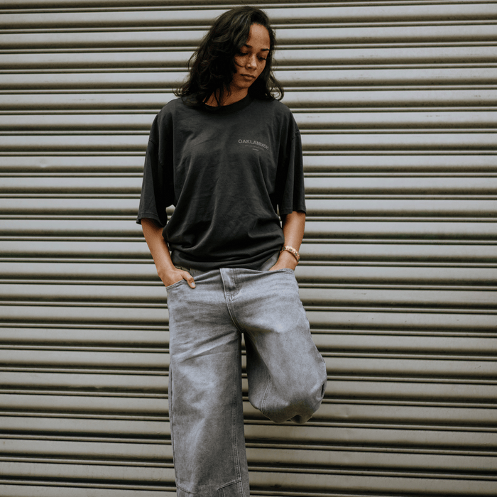 Against a metal roll-up door, a person wears the Oaklandish Modern Type Heavy Tee—a faded black, boxy fit with "Oakland" on it—and light gray jeans. Their hands are in pockets, medium-length dark hair frames their calm expression as they look downwards, suggesting an urban vibe.