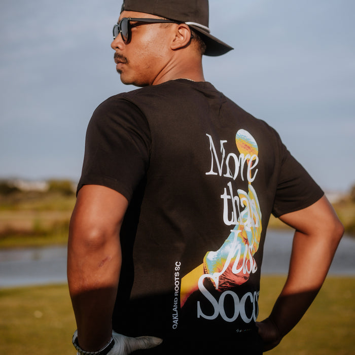 A man wearing an Oakland Roots SC "More Than Soccer Tee" with a graphic of a soccer player on the back stands outdoors. He sports an Oaklandish Classic fit t-shirt, a backwards baseball cap, sunglasses, and white gloves. The background features a grassy area and a body of water under a clear sky.