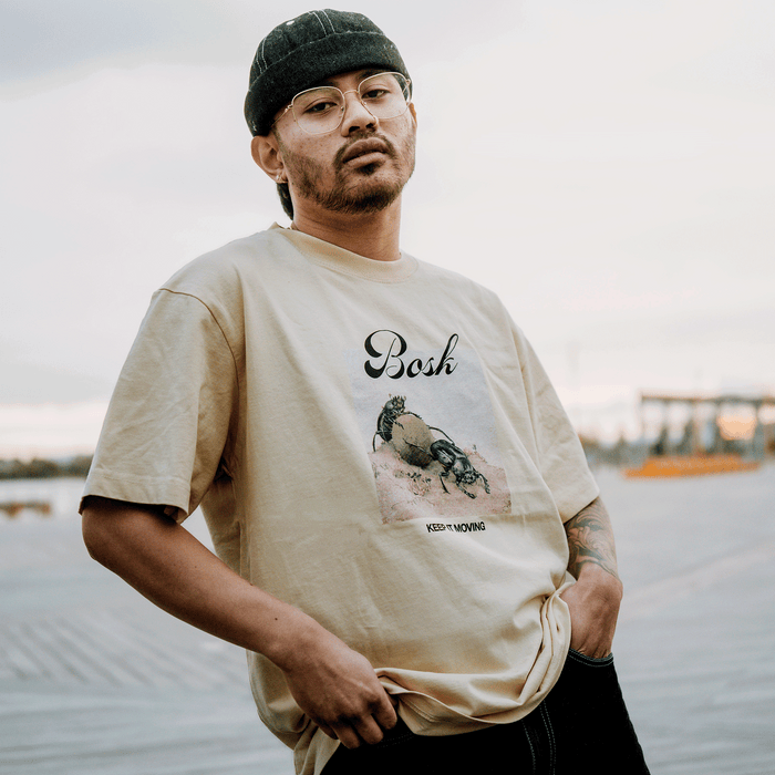 A man in glasses and a black cap stands on a boardwalk, wearing the BOSK Moving Tee—a classic fit, heavy-weight beige T-shirt with a crab graphic and "KEEP MOVING." He looks at the camera, holding his 100% cotton shirt. Background is blurred.