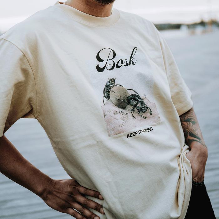 A person wears the BOSK Moving Tee, a classic fit light beige heavy-weight cotton shirt with two beetles on a rock, “Bosh” script above, and “KEEP THINKING” in all caps below. Hands rest on hips outdoors.