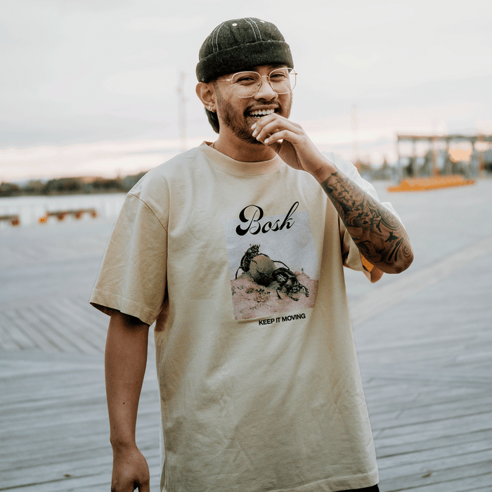 A smiling person in glasses and a dark beanie stands on a boardwalk at sunset, wearing the BOSK Moving Tee— a classic fit heavy-weight cotton T-shirt with a crab graphic and “Bosh” and “Keep It Moving” text. Tattoos show on their right forearm.
