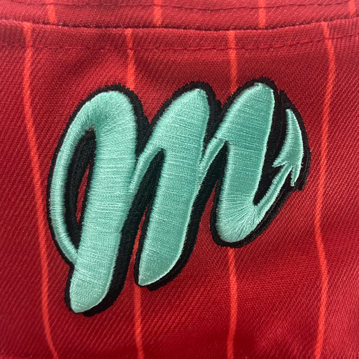 Here’s a rewritten version of the sentence using your product data: Close-up of the New Era Diablos Paisley Bucket by Other, showcasing vivid red polyester fabric with fine white pinstripes and an embroidered mint green “M” outlined in black. The letter finishes with a devil tail on the right, combining sporty flair with playful, devilish character—an ideal statement detail for this distinctive bucket hat.