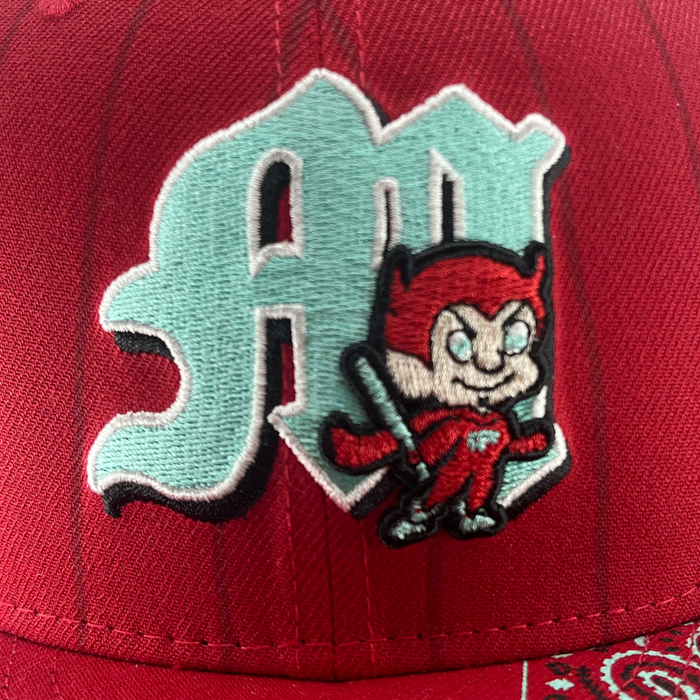 A close-up of the New Era Diablos Pinstripe 59FIFTY Cap by Other, showcasing an embroidered light blue “M” on the red fitted crown. In front of the letter stands a cartoon red devil holding a bat and dressed in a light blue outfit. The cap also features a partial paisley pattern along the bottom edge.