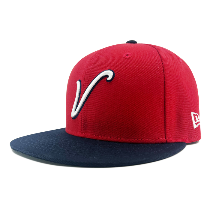 A red New Era El Aguila Alt 59FIFTY Cap featuring a navy blue flat brim. The front showcases a large, white, stylized “V” outlined in navy, while the white New Era logo is embroidered on the side. This fitted cap is displayed against a plain white background.