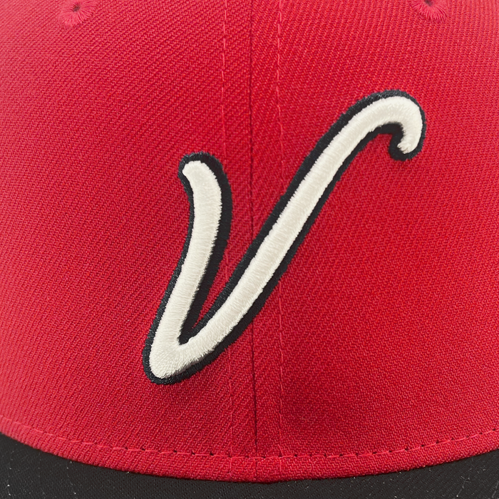 A close-up of the New Era El Aguila Alt 59FIFTY Cap from Other showcases a red cap with a black bill, featuring a white, stylized “V” outlined in black centered on the front panel. The red fabric displays a visible weave texture and includes ventilation eyelets near the top. This fitted 59FIFTY design blends classic style with comfort and quality craftsmanship.