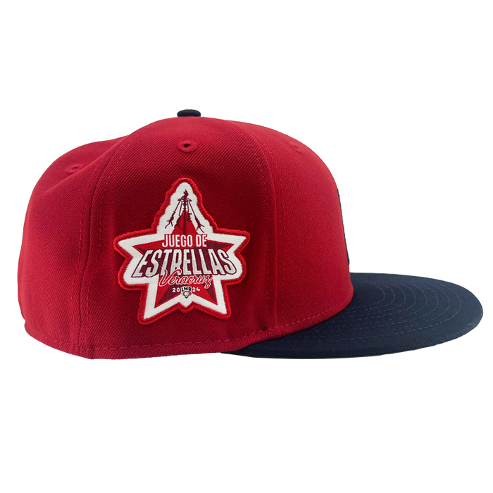 New Era El Aguila Alt 59FIFTY Cap by Other, featuring a red crown with a navy blue flat brim. The side showcases a star-shaped patch with white and red text that reads “Juego de Estrellas Uruguay” and “2023,” alongside a graphic of a tree and mountain. This fitted cap is set against a plain white background.