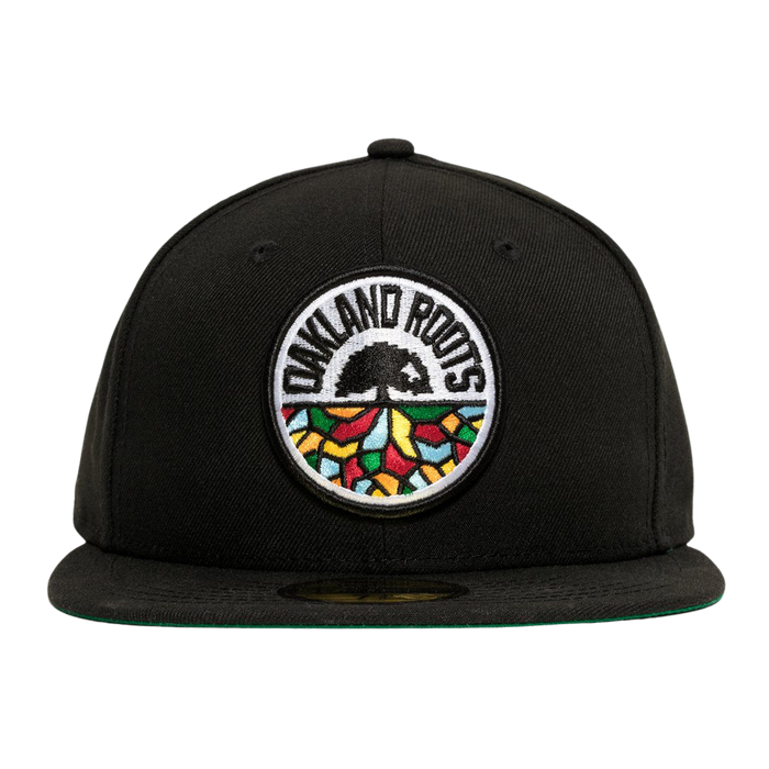A New Era Oakland Roots SC 59FIFTY Fitted Cap in black, featuring a circular embroidered patch on the front. The patch showcases the words "Oakland Roots" at the top, a black tree in the center, and colorful geometric shapes resembling a mosaic at the bottom. This sports club cap comes with a flat brim and a green underside.