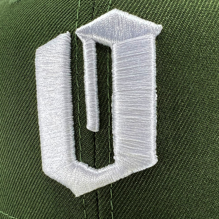 Close-up of a green New Era Official 59FIFTY Fitted Cap by Other, featuring a large, raised white embroidered symbol with a stylized angular design and vertical bar on the left. Fine stitching detail is visible on both the logo and the 59FIFTY cap itself.
