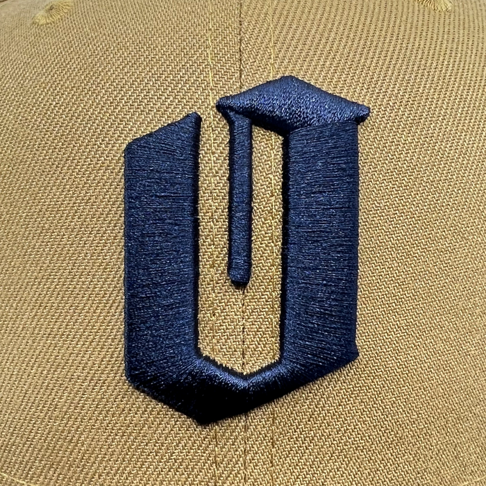 A close-up of a tan New Era Official 59FIFTY Fitted Cap by Other highlights a large, navy blue, embroidered geometric “U” logo centered on the front. The prominent stitching and detailed logo stand out against the textured fabric, reflecting the signature quality of New Era’s 59FIFTY design.