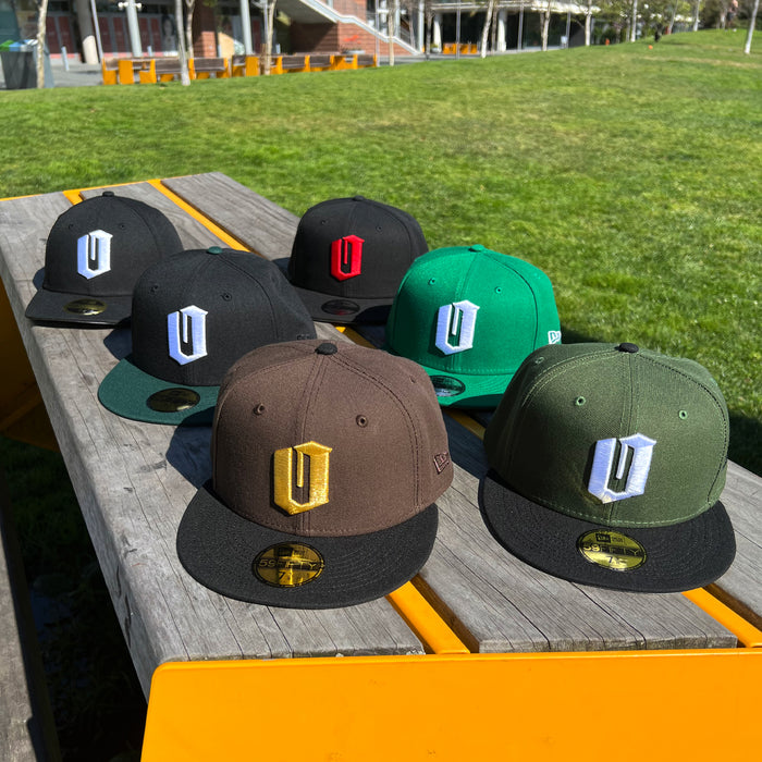 Six flat-brim New Era Official 59FIFTY Fitted Caps by Other, each featuring a large embroidered “U” logo in various colors, are displayed on a wooden bench outdoors. These caps come in black, brown, green, and olive colorways with assorted logo variations. The background features grassy outdoor scenery and seating.